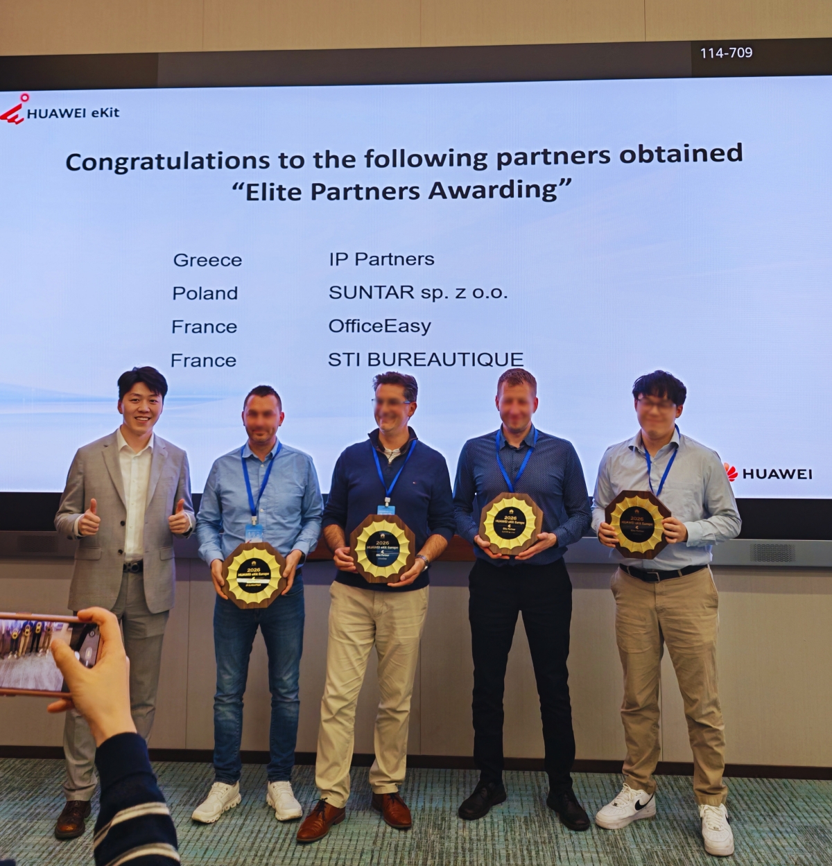 Group photo of the 2026 HUAWEI eKit Europe Elite Partners — including IP Partners from Greece, SUNTAR from Poland, OfficeEasy and STI BUREAUTIQUE from France — holding their plaques in front of the announcement screen at Huawei HQ.